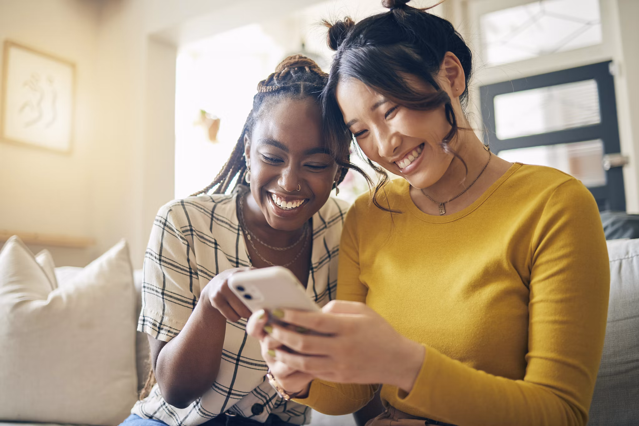 Two women smile and laugh while looking at a smartphone, sitting on the couch together.