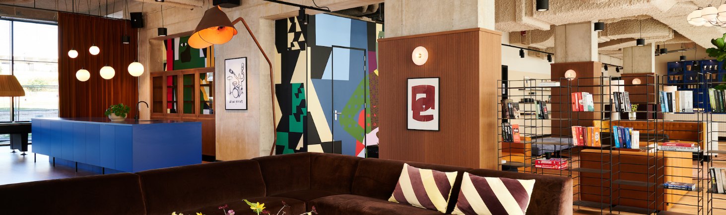 Modern cozy interior of a lounge with eclectic decor, featuring a large brown sofa, colorful abstract art, and a well-stocked bookshelf.