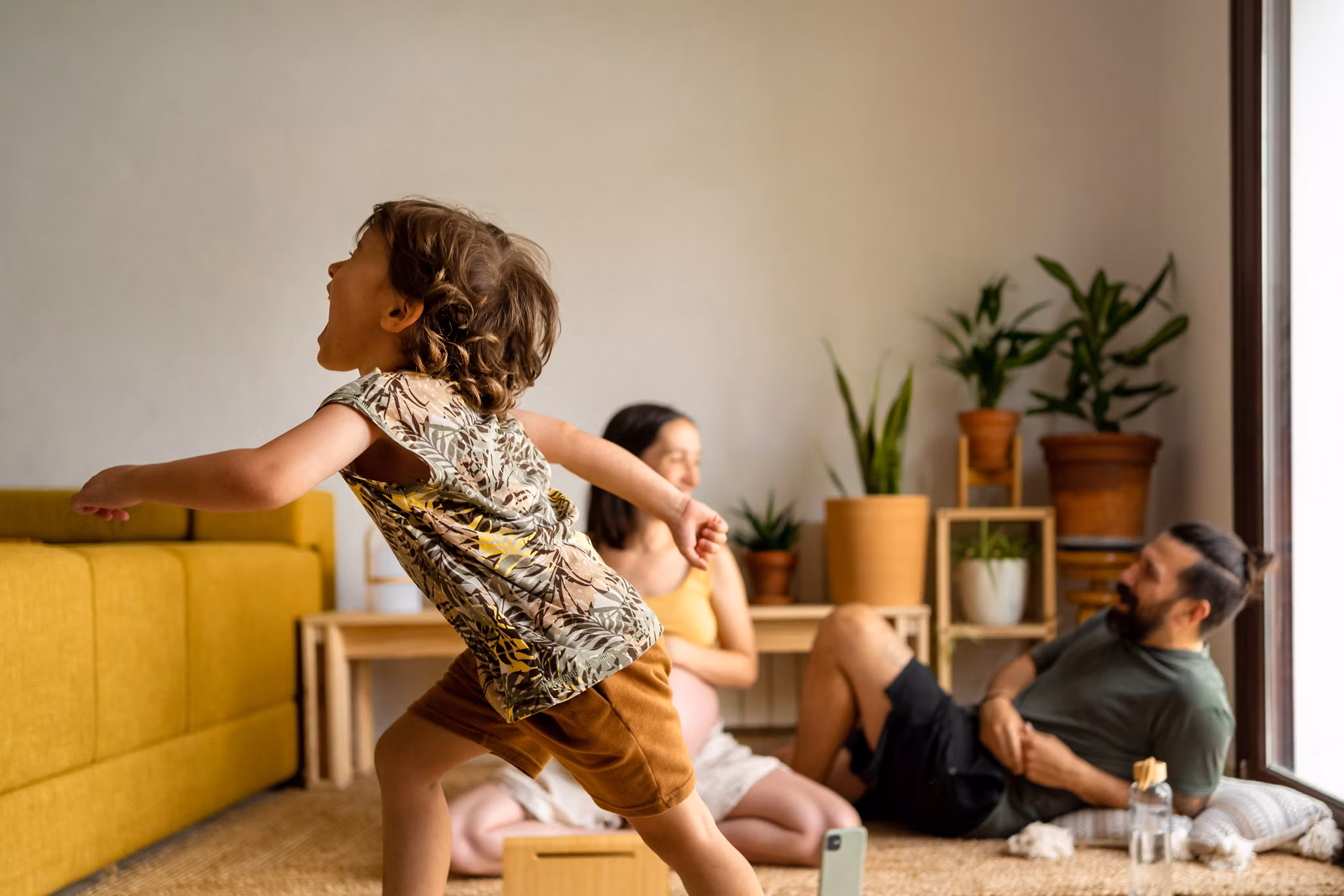A child running in a room while the parents are sitting on the floor talking and laughing.